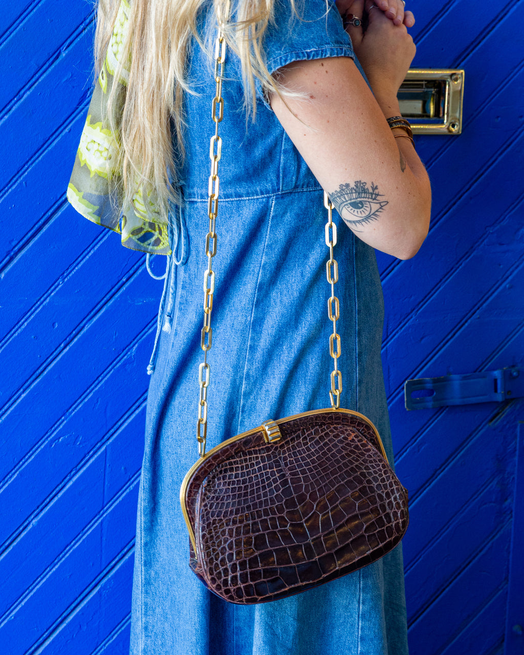 Three-quarter view of a vintage brown alligator handbag with a gold-tone frame, worn crossbody on a gold chain over a blue denim dress against a blue door.