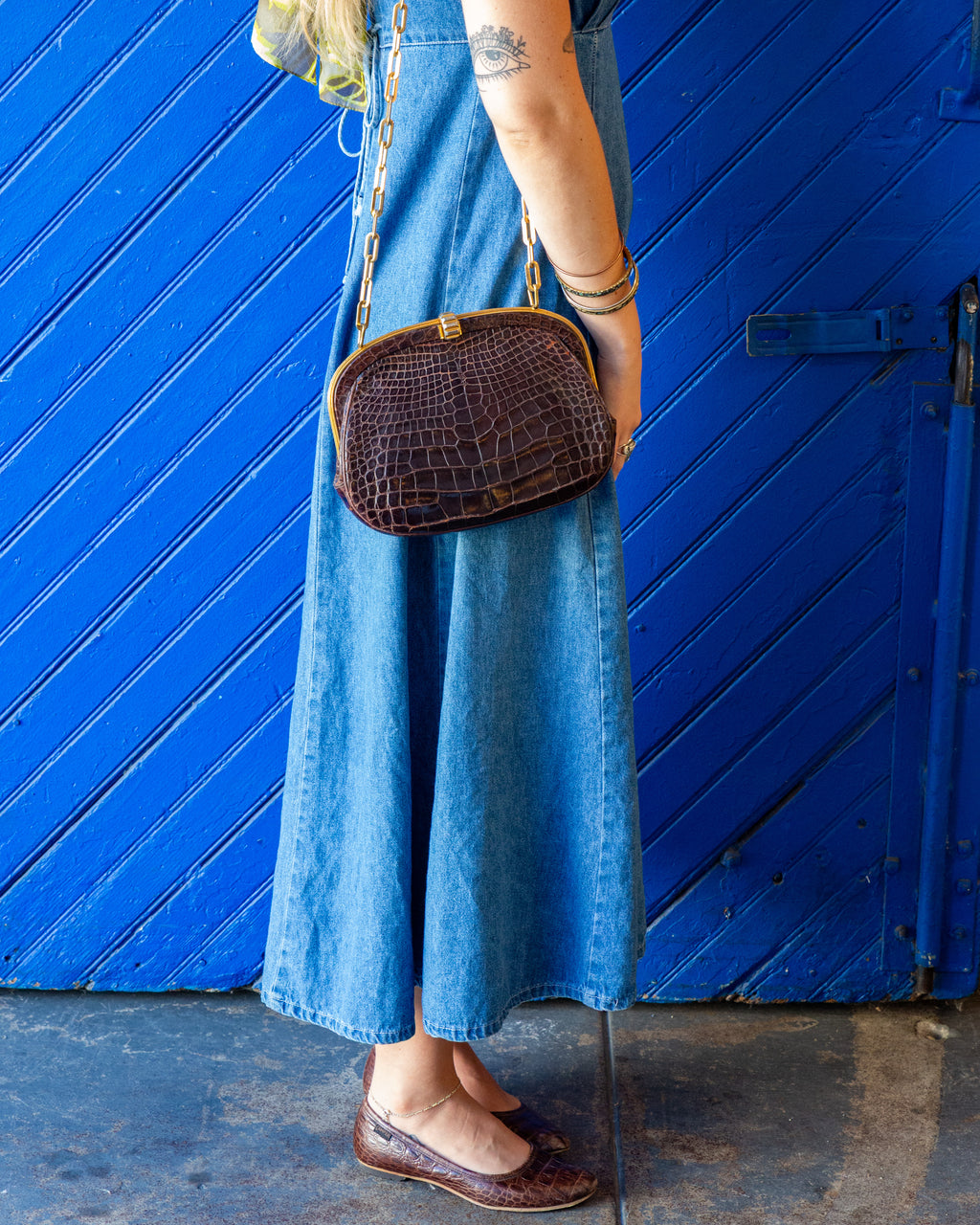 Full-length view of a vintage brown alligator handbag resting at the hip, worn crossbody on a gold chain over a blue denim dress.