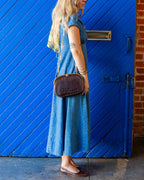 Side profile of a vintage brown alligator handbag worn crossbody on a gold chain over a blue denim dress, in front of a blue door and brick wall.