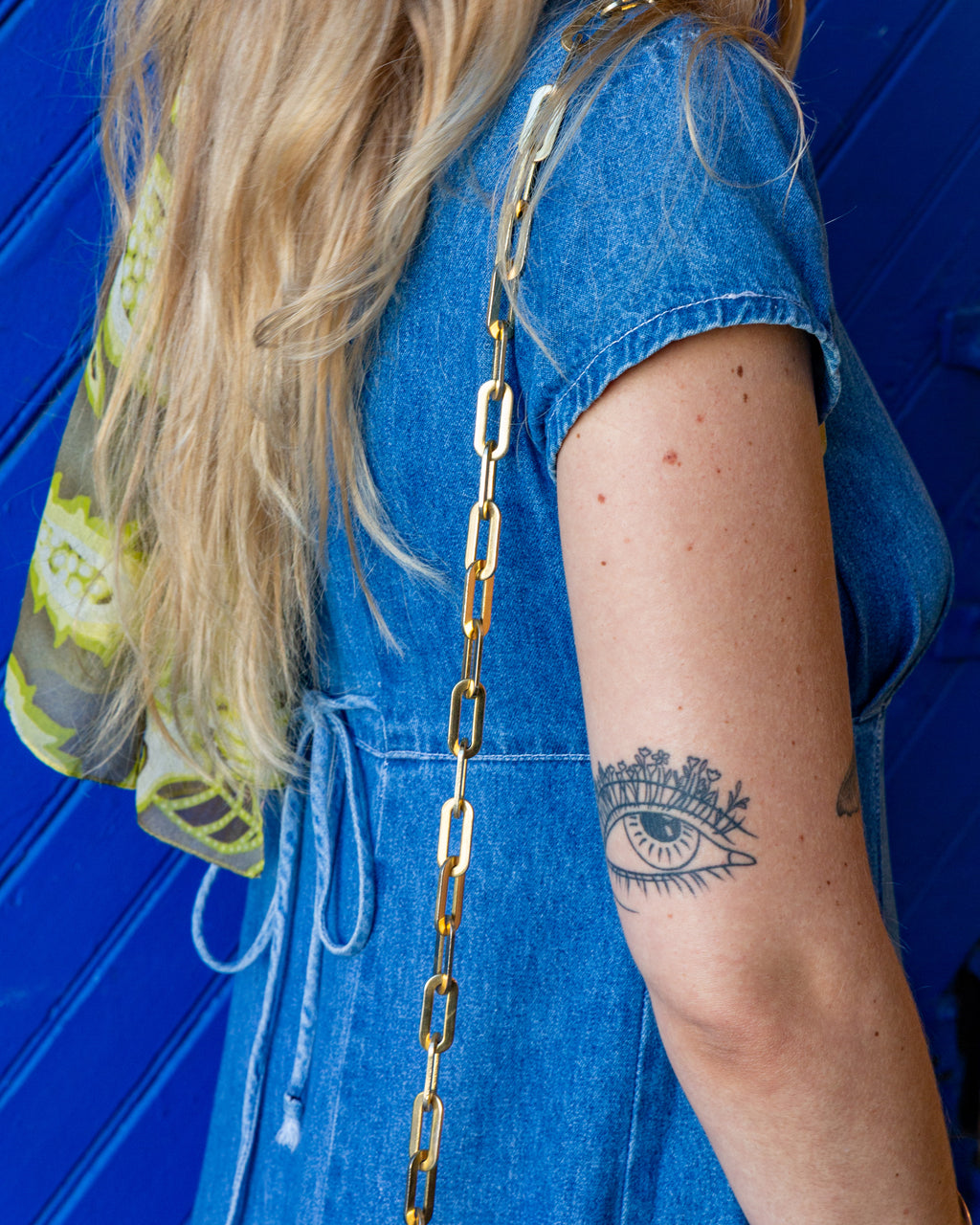 Close view of the flat gold-tone paperclip chain strap of a vintage brown alligator handbag worn over a blue denim dress.