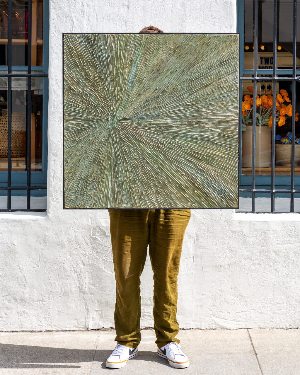 Square abstract painting in sage and ochre impasto with a radiating starburst pattern, held straight on against a white stucco storefront wall.