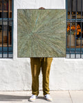 Square abstract painting in sage and ochre impasto with a radiating starburst pattern, held straight on against a white stucco storefront wall.
