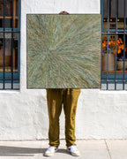 Square abstract painting in sage and ochre impasto with a radiating starburst pattern, held straight on against a white stucco storefront wall.