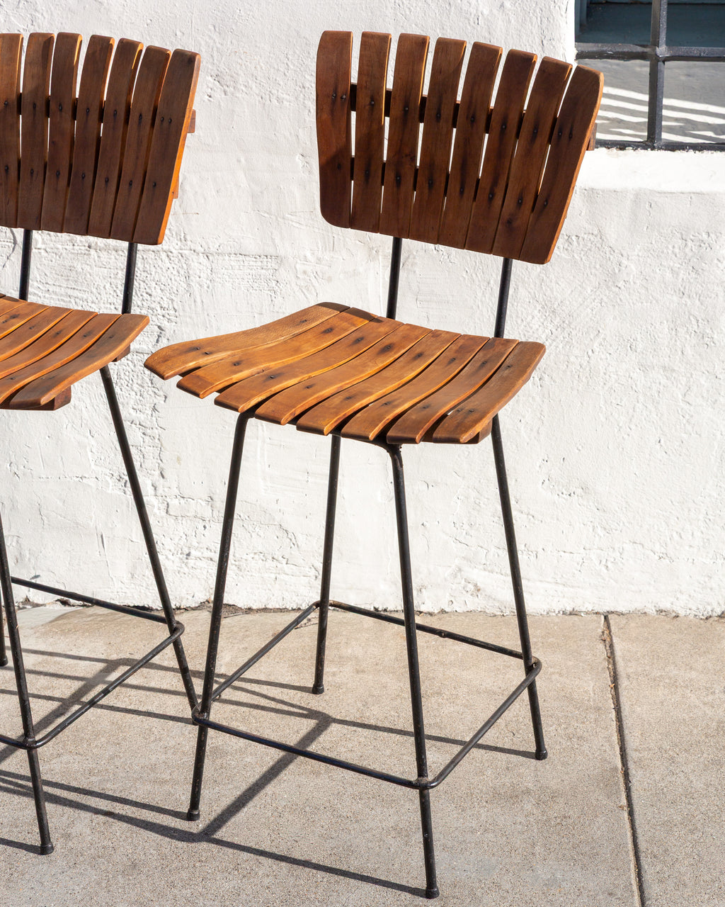 Three-quarter view of a pair of mid century bar stools with slatted wood seats and backs on black metal frames against a white stucco wall