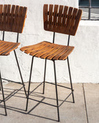 Three-quarter view of a pair of mid century bar stools with slatted wood seats and backs on black metal frames against a white stucco wall