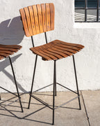 Full view of an Arthur Umanoff style bar stool with curved wood slat seat and back and black rod legs with footrest