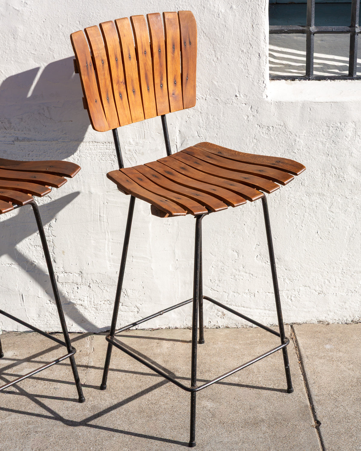 Full view of an Arthur Umanoff style bar stool with curved wood slat seat and back and black rod legs with footrest