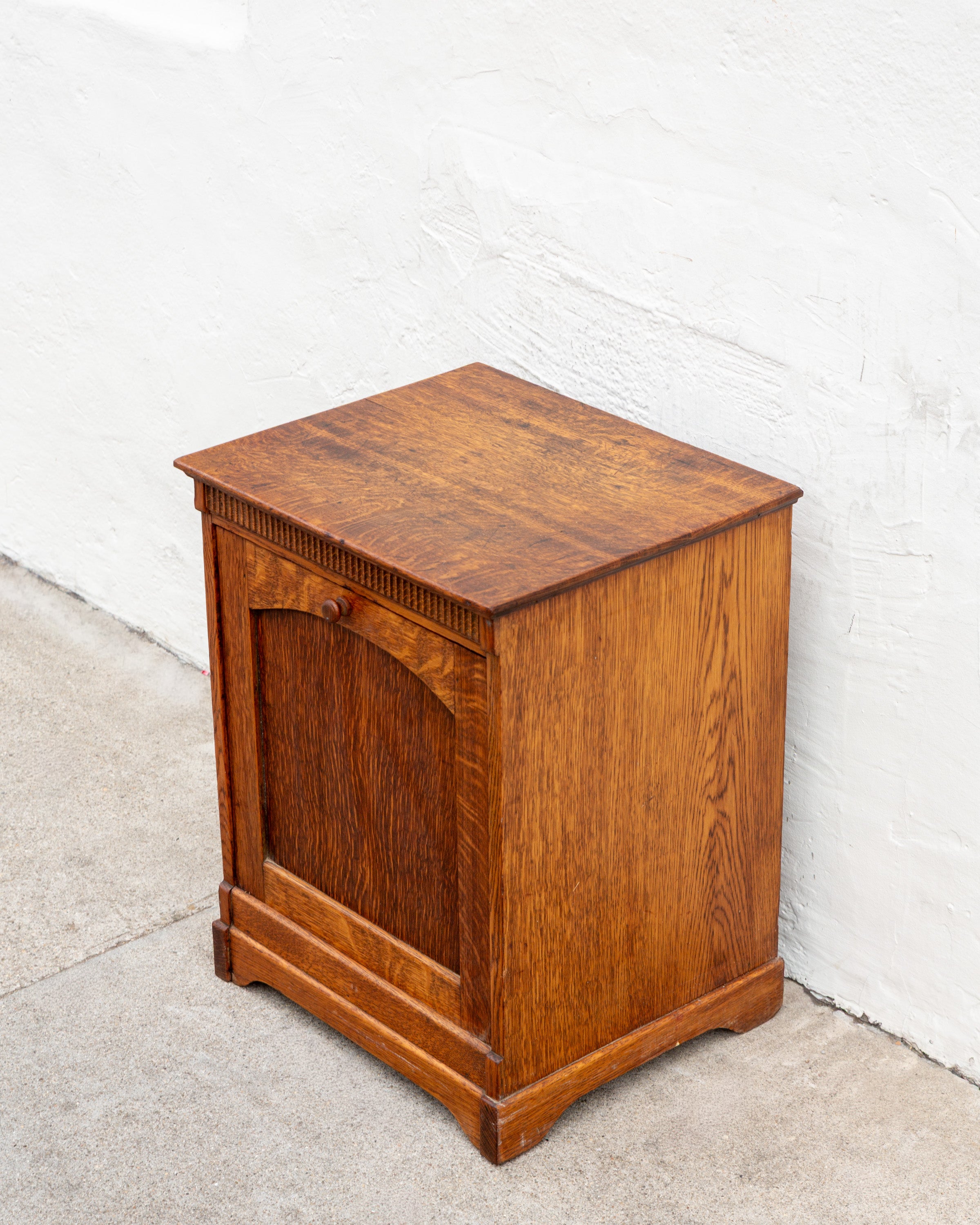 Three-quarter view of an antique tiger oak tilt front bin cabinet in warm golden brown against a white stucco wall.