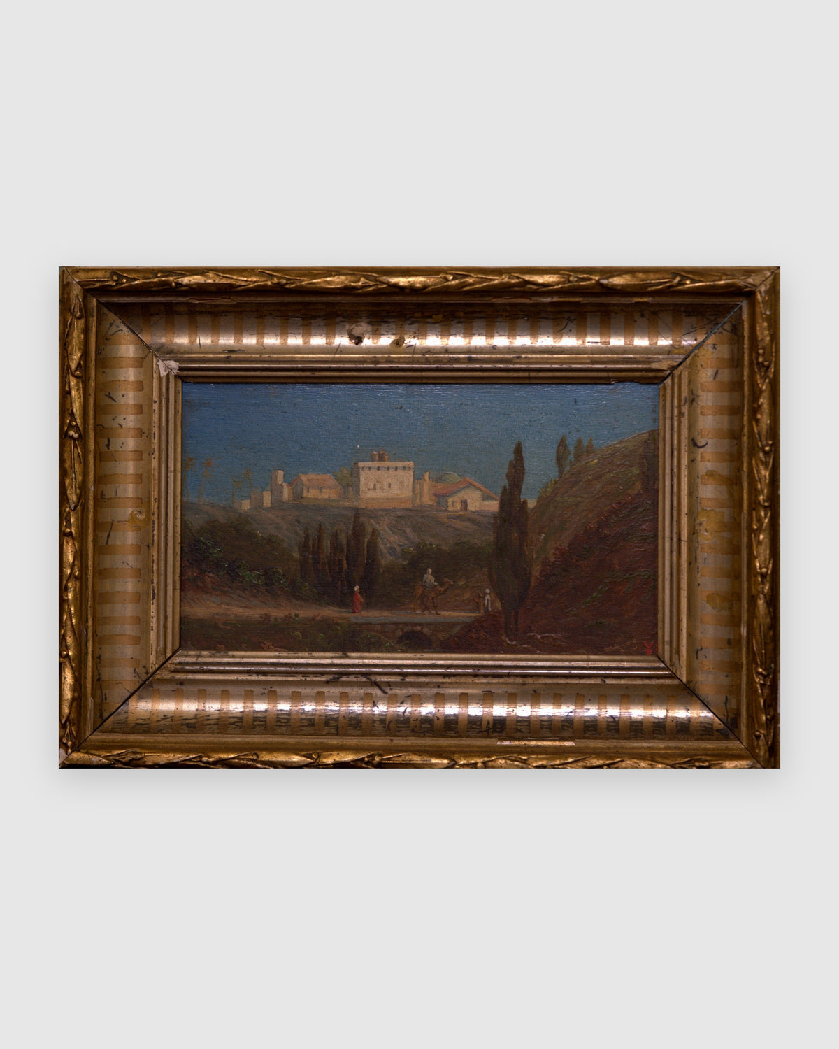 Straight-on view of a small Italian landscape of a hilltop villa and cypresses in a carved gilt and silver frame, on a light gray background.