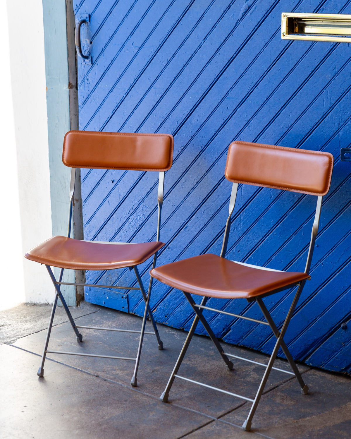 Three-quarter view of a pair of folding chairs with cognac leather seats and backs on gray metal X-frames against a blue painted door.