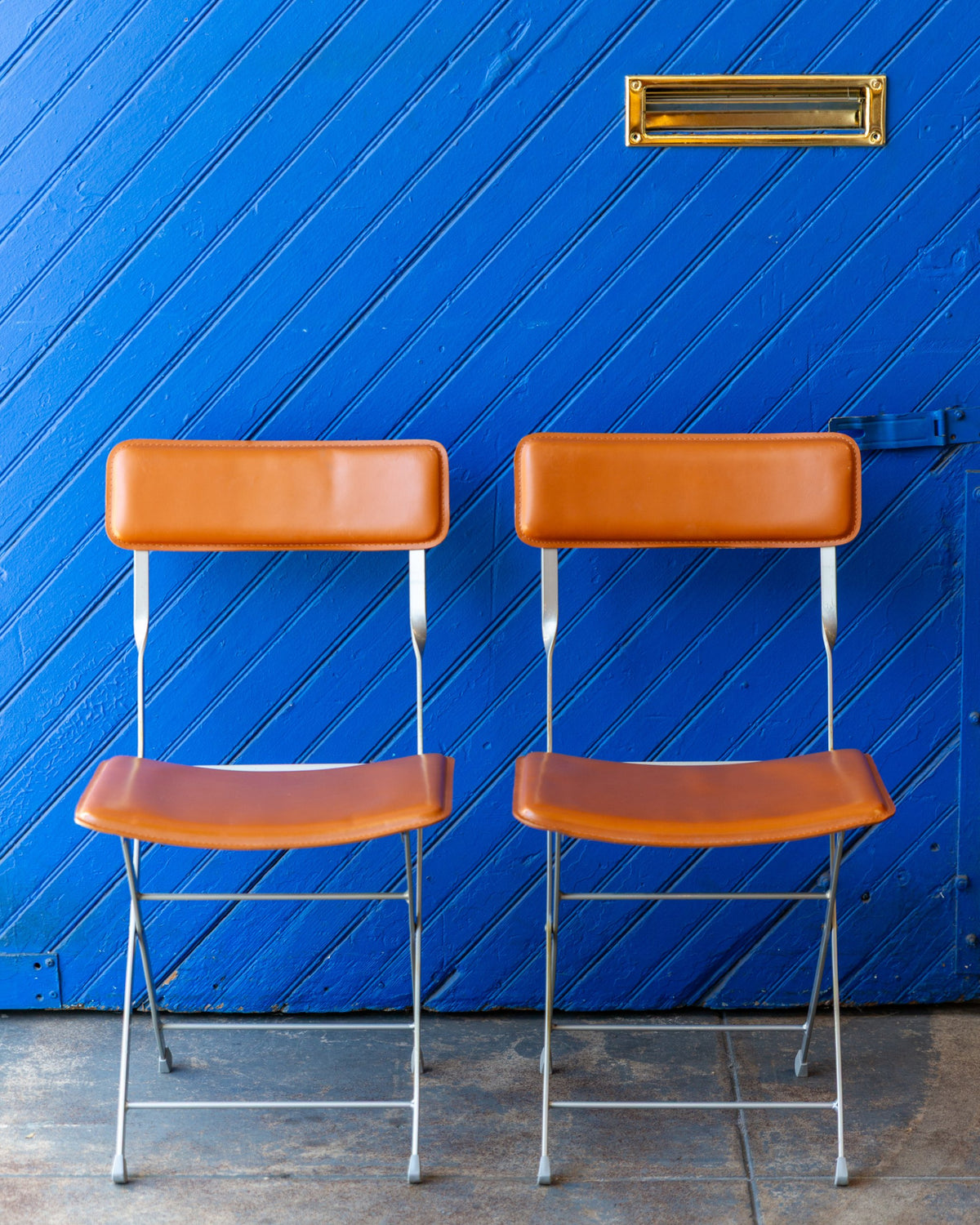 Front view of a pair of cognac leather and gray metal folding chairs side by side against a blue door.