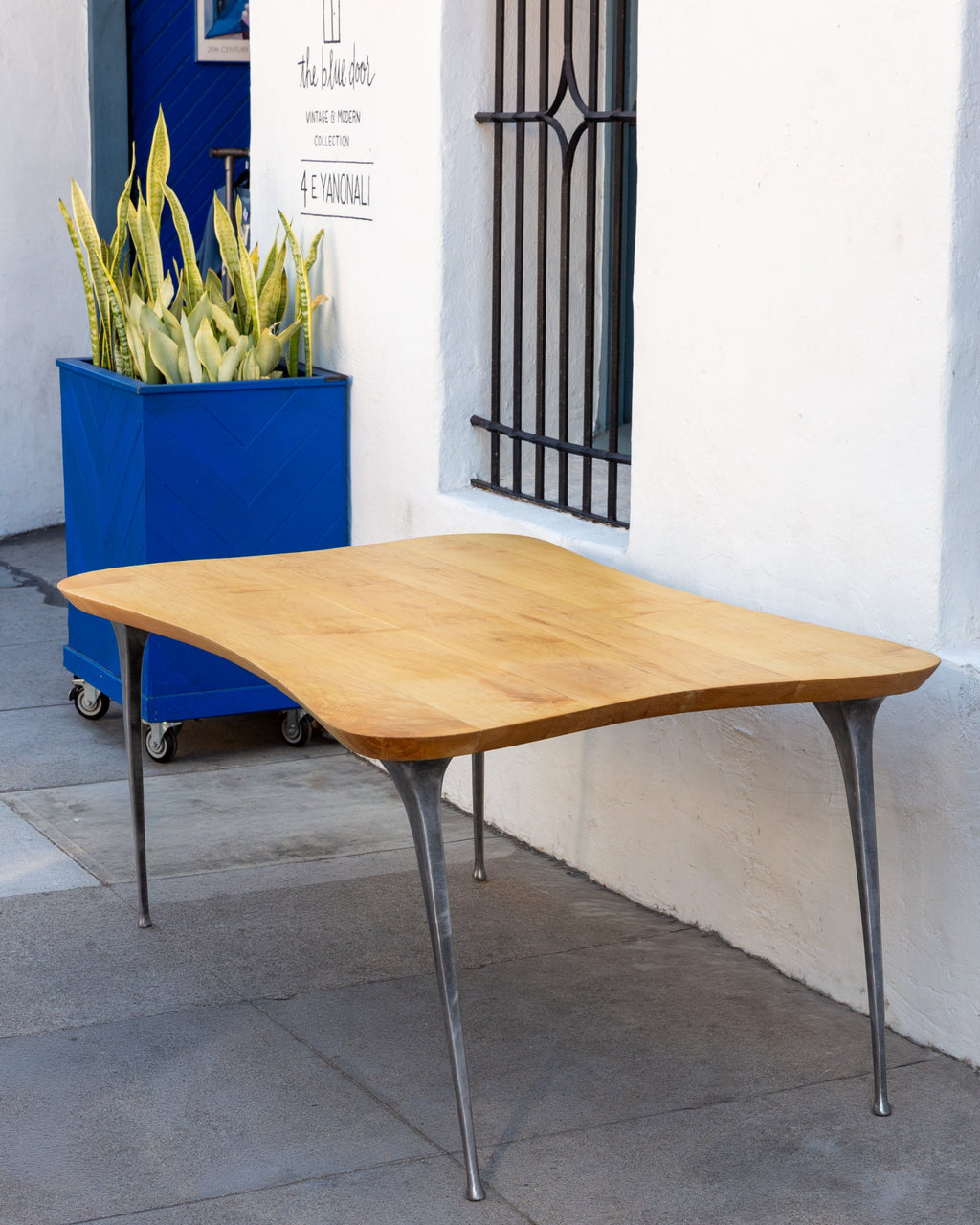 Three-quarter view of a 1970s freeform solid maple table on four tapered cast aluminum legs against a white stucco wall.