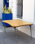 Three-quarter view of a 1970s freeform solid maple table on four tapered cast aluminum legs against a white stucco wall.