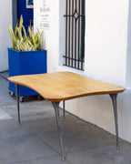 Three-quarter view of a 1970s freeform solid maple table on four tapered cast aluminum legs against a white stucco wall.