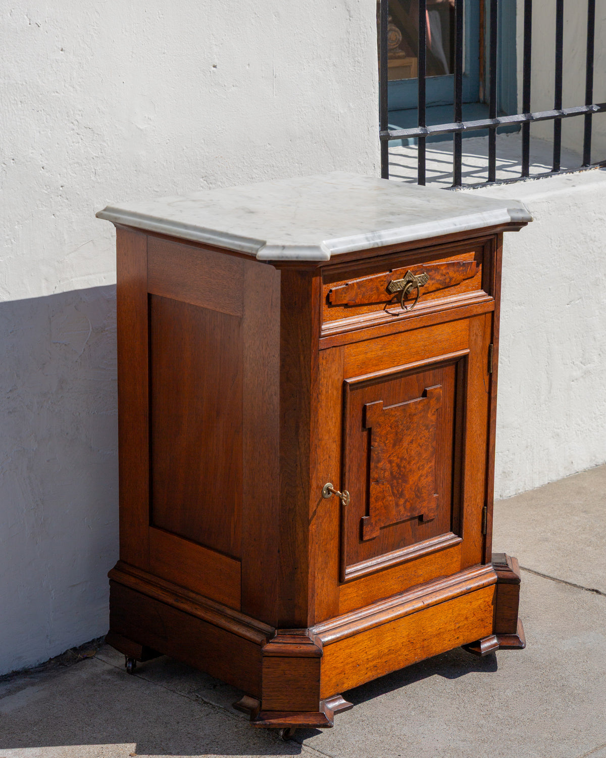 Three-quarter view of an antique Victorian walnut cabinet with a white marble top, single drawer and paneled cupboard door, against a white stucco wall