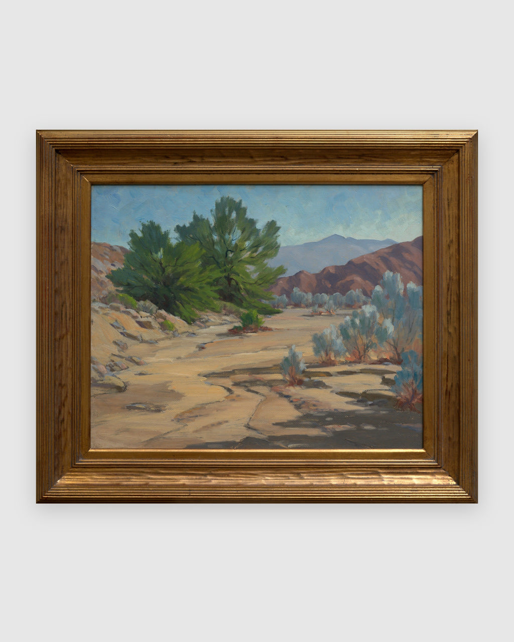 Straight-on view of a Jack McCartney desert landscape with a green smoke tree, sage and rose hills in a stepped gilt frame, on a light gray background.