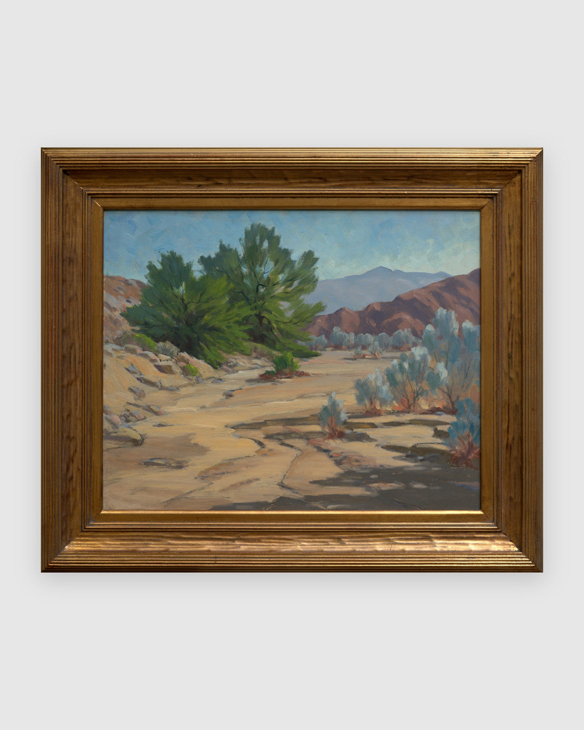 Straight-on view of a Jack McCartney desert landscape with a green smoke tree, sage and rose hills in a stepped gilt frame, on a light gray background.