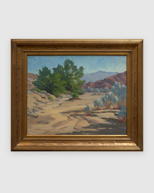 Straight-on view of a Jack McCartney desert landscape with a green smoke tree, sage and rose hills in a stepped gilt frame, on a light gray background.