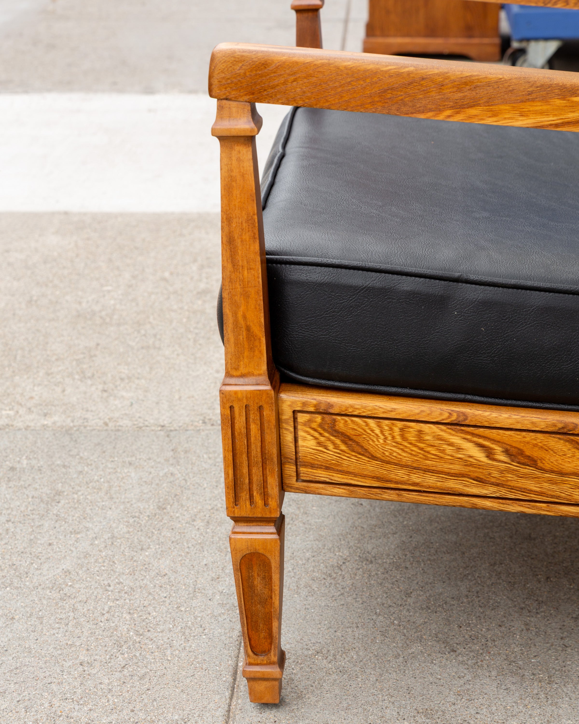 Detail of the fluted, tapered front leg and arm post on a refinished golden oak arm chair, with the edge of the black cushion behind
