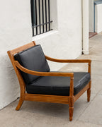 Three-quarter view of a mid century oak arm chair with black cushions against a white stucco wall