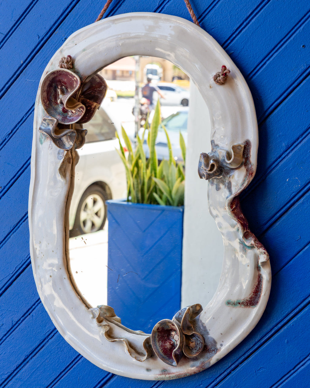 Three-quarter view of a vintage white glazed ceramic wall mirror with applied red and blue flowers, hanging on a blue plank wall.