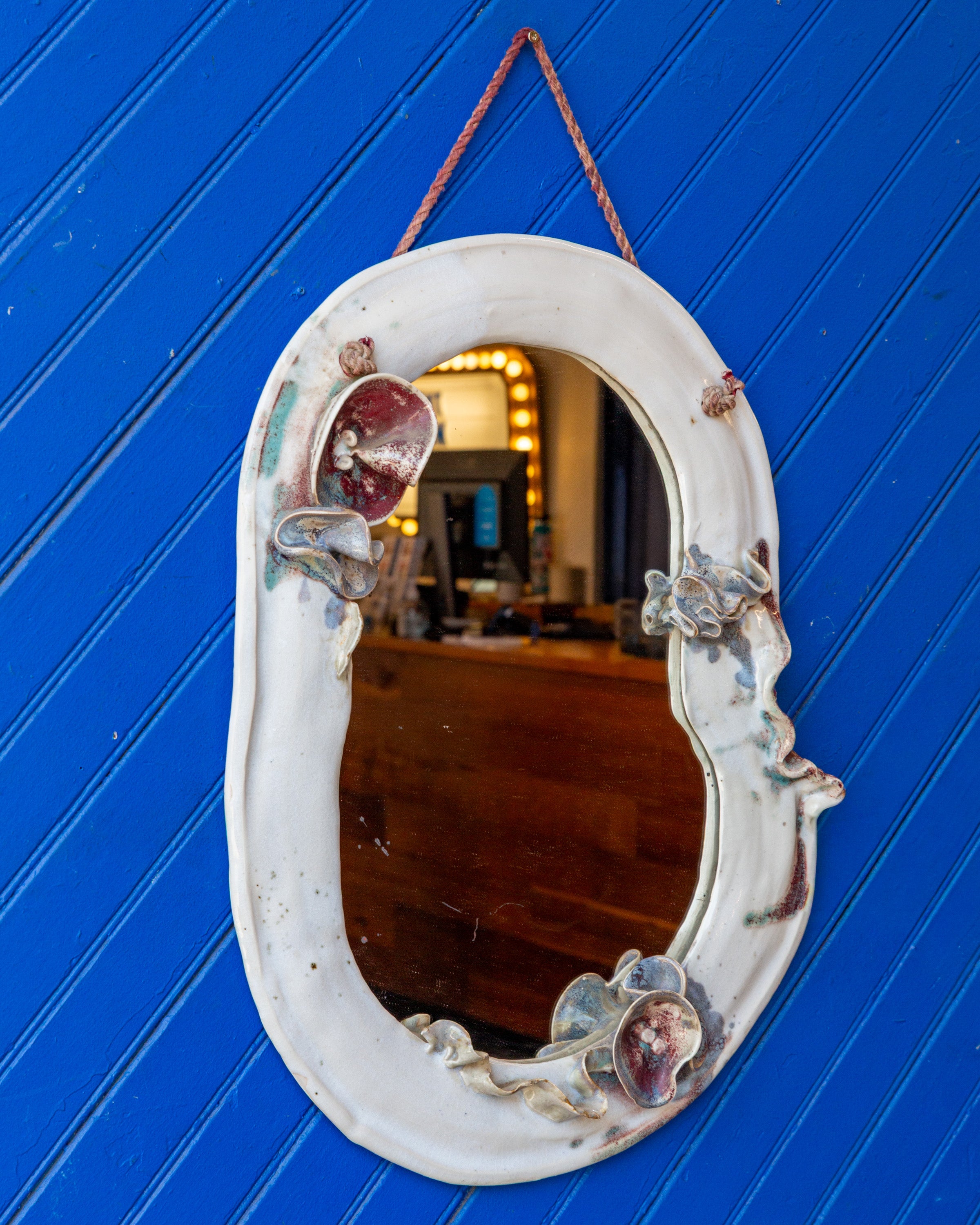 Front view of a vintage white ceramic mirror with sculpted flowers on its wavy frame, hanging on a blue plank wall.