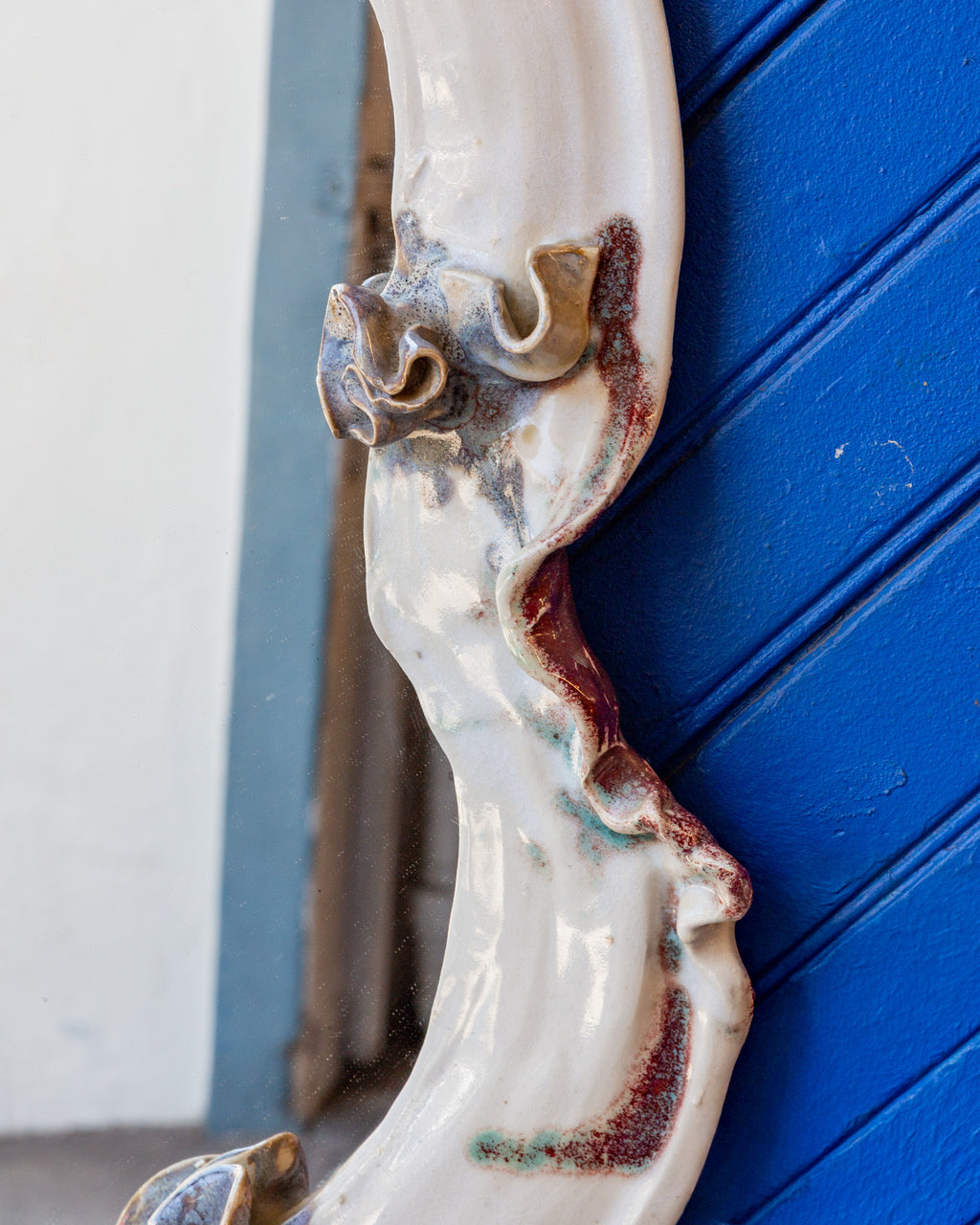 Angled view of the right side of the white ceramic mirror frame, showing ruffled clay edges glazed in oxblood and green.