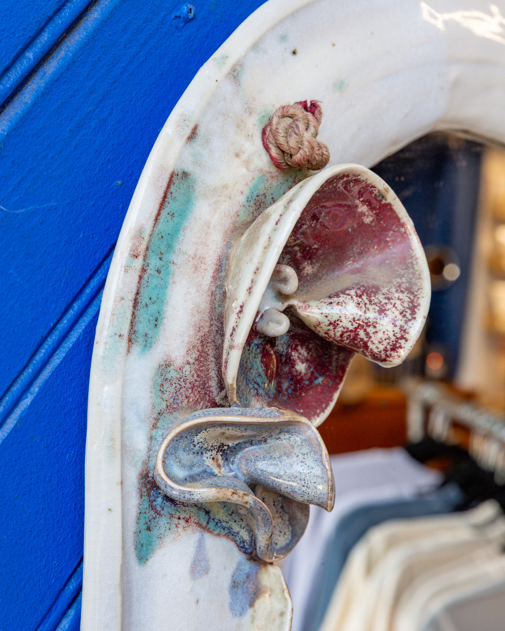 Tight detail of a deep red ceramic flower and knotted cord on the white glazed mirror frame.