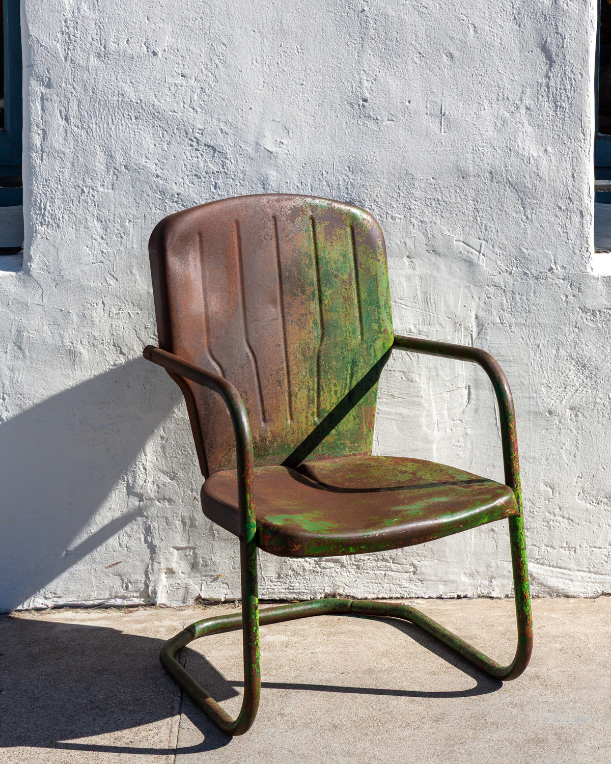 Three-quarter view of a 1950s steel motel chair with worn green paint and rust patina against a white stucco wall