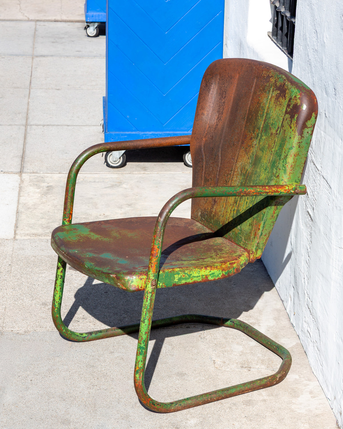 Front three-quarter view of a steel motel chair with heavy green and rust patina beside a blue bin