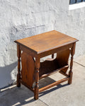 Three-quarter view of a solid oak side table with turned legs and a trough book shelf against a white stucco wall
