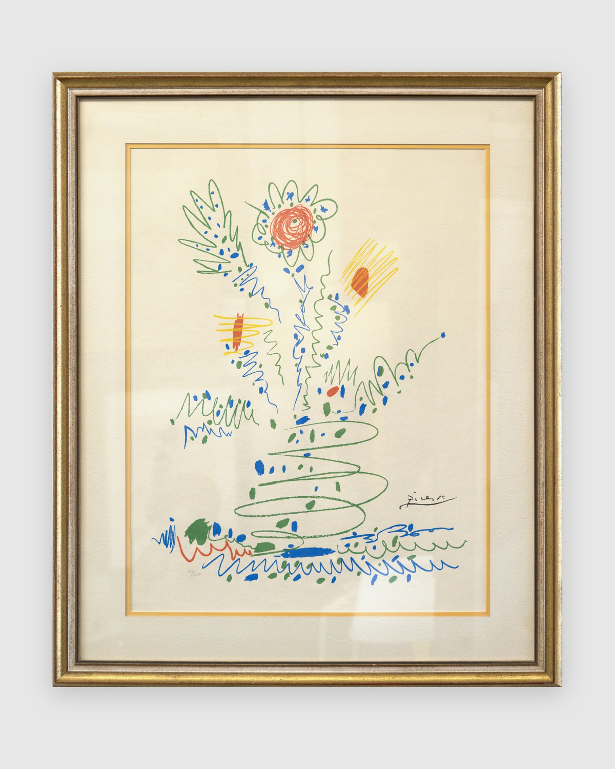 Straight-on view of a framed Picasso color lithograph of a sprouting flower bouquet in green, blue, red and yellow, cream mat with gold fillet, gilt and silver frame, on a light gray background.