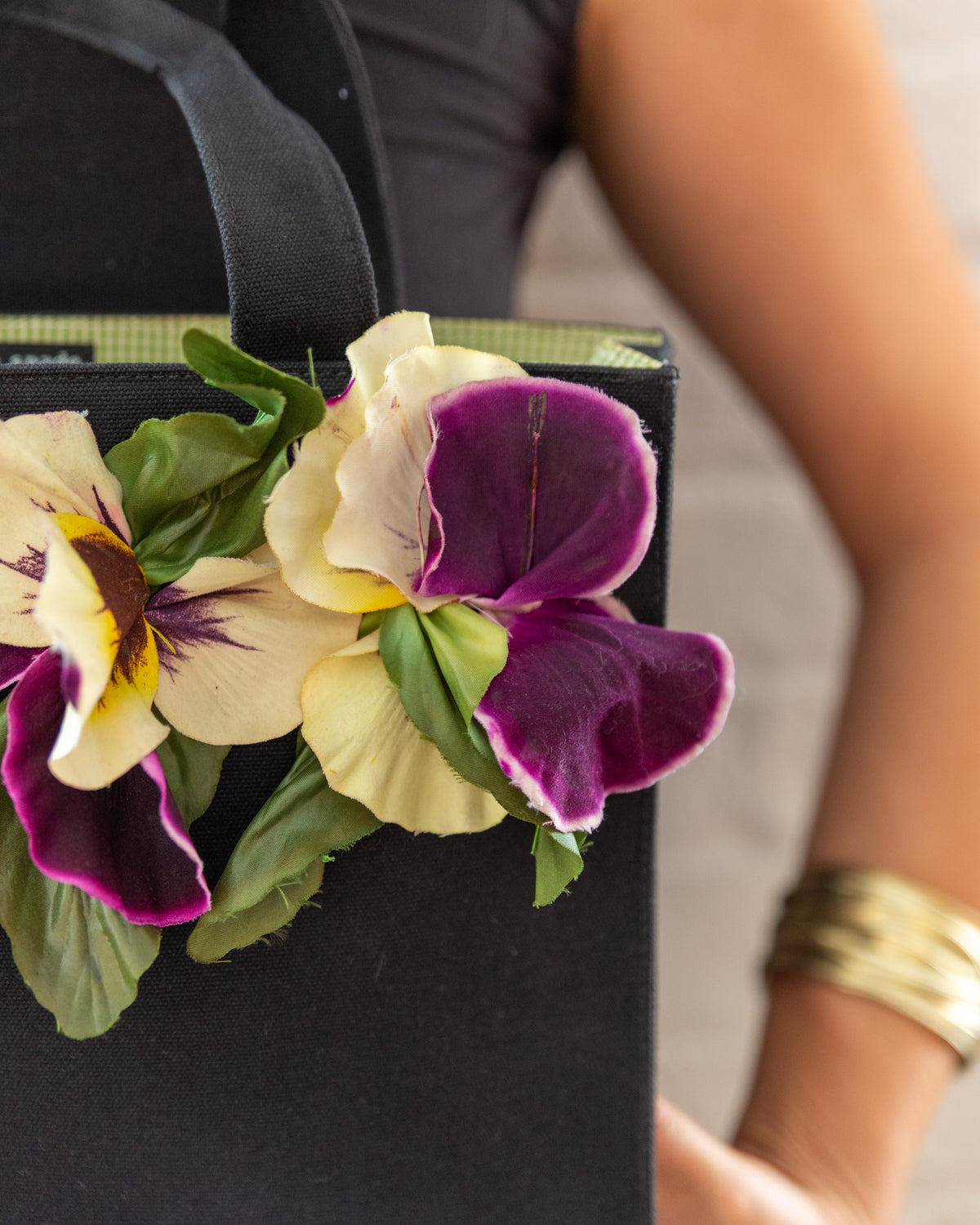Angled close view of the fabric pansies at the top corner of the black canvas tote, with frayed petal edges visible.