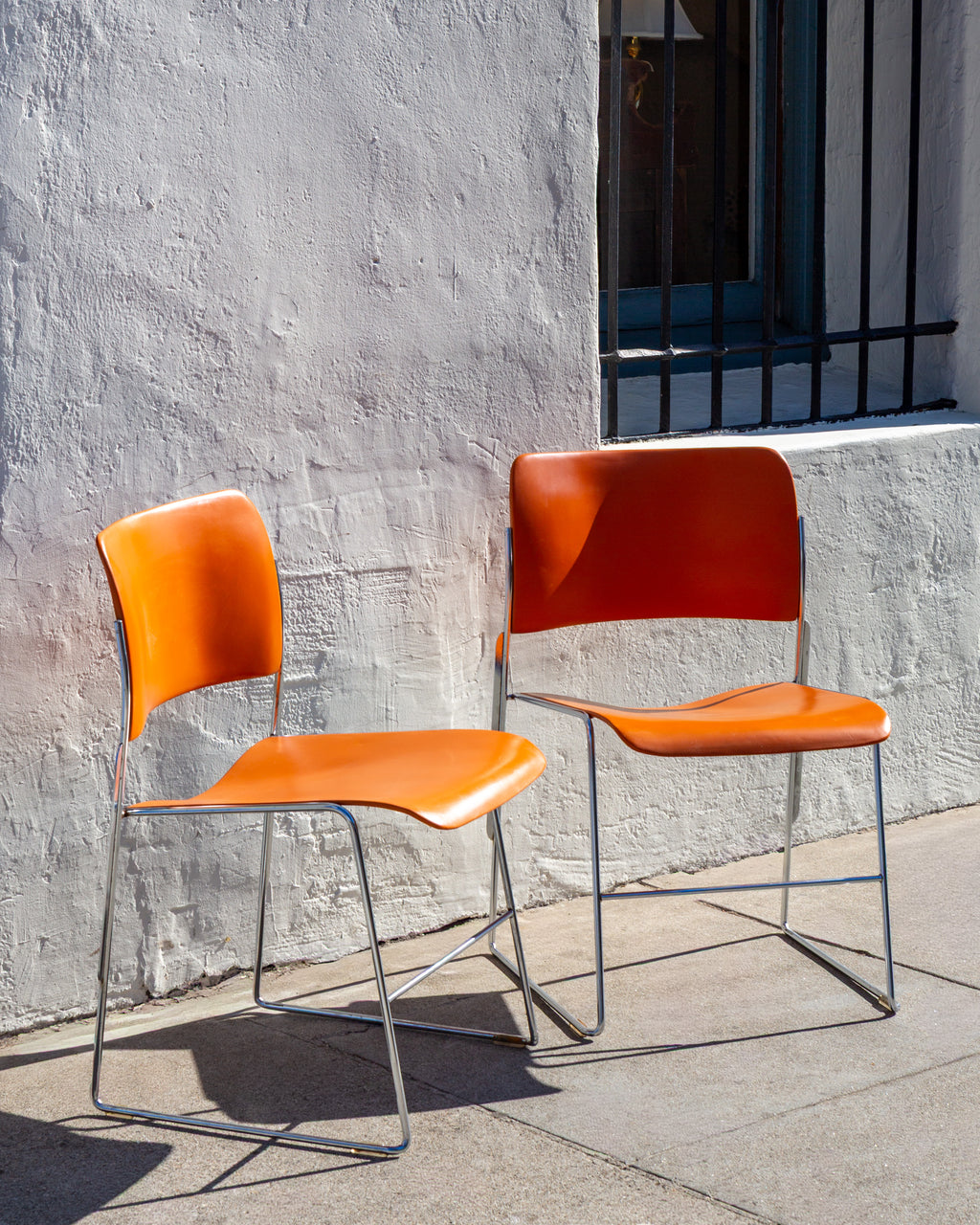 Three-quarter view of a pair of David Rowland 40/4 stacking chairs with orange seats and chrome frames against a white stucco wall
