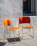 Three-quarter view of a pair of David Rowland 40/4 stacking chairs with orange seats and chrome frames against a white stucco wall