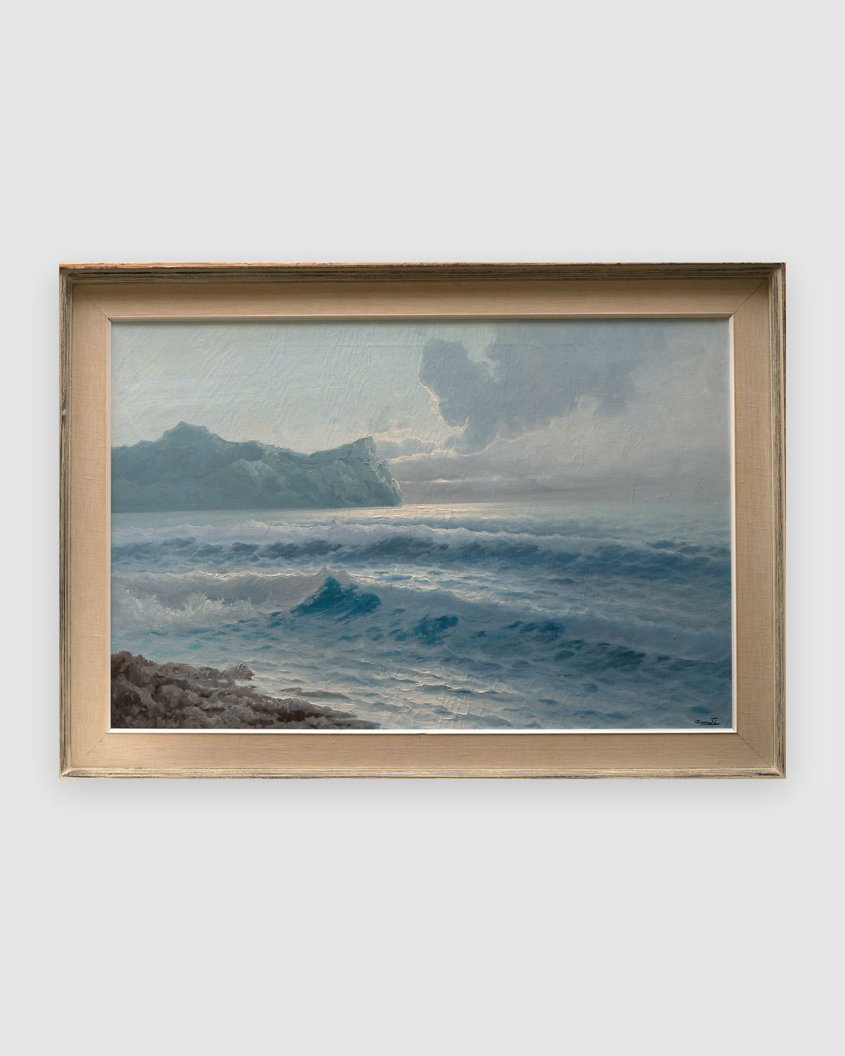 Straight-on view of a 1960s seascape of turquoise breaking waves, rocks and a misty headland in a whitewashed frame with linen liner, on a light gray background.