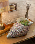Three-quarter view of a conch-shaped planter covered in small pink and lavender shells, planted with green succulent, on a copper-topped table