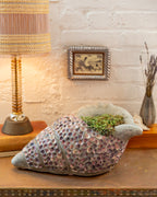 Front three-quarter view of a gray conch planter inlaid with pink shells in spiral bands beside a lamp