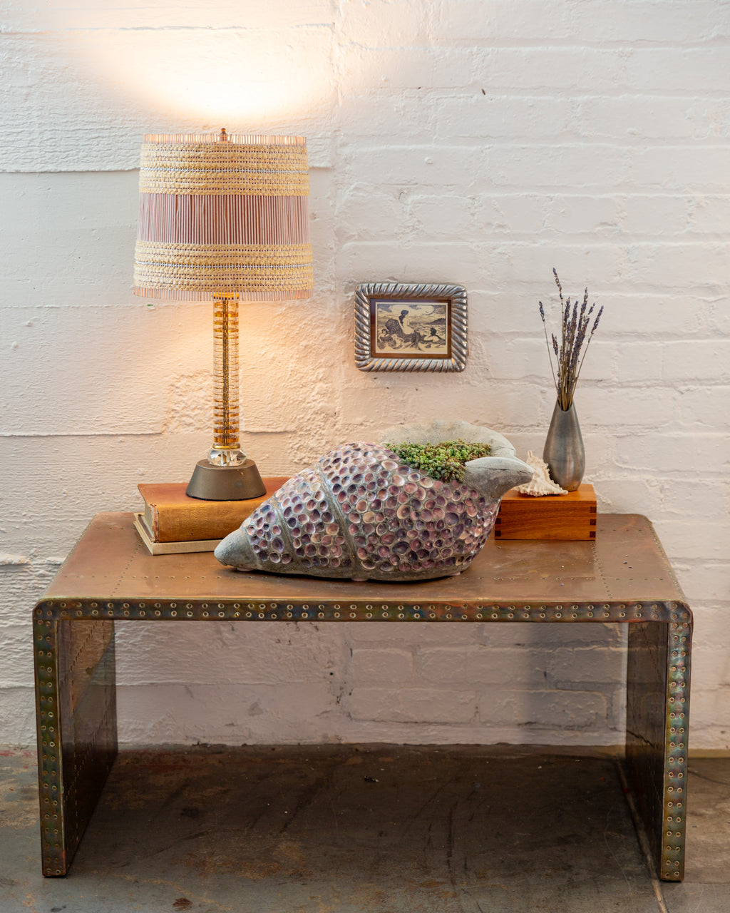 Shell mosaic conch planter styled on a low table against a white brick wall with a lamp and small framed print