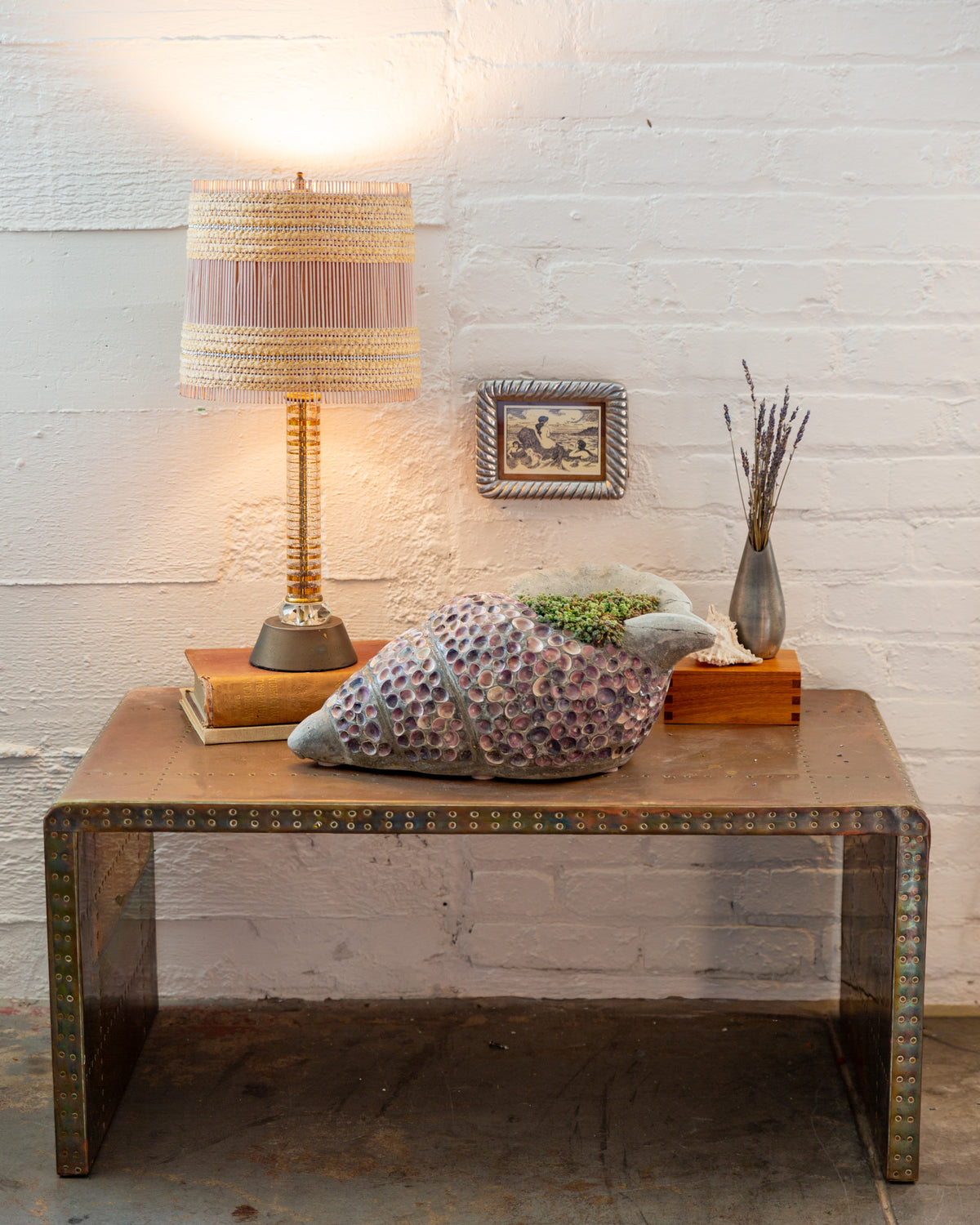 Shell mosaic conch planter styled on a low table against a white brick wall with a lamp and small framed print