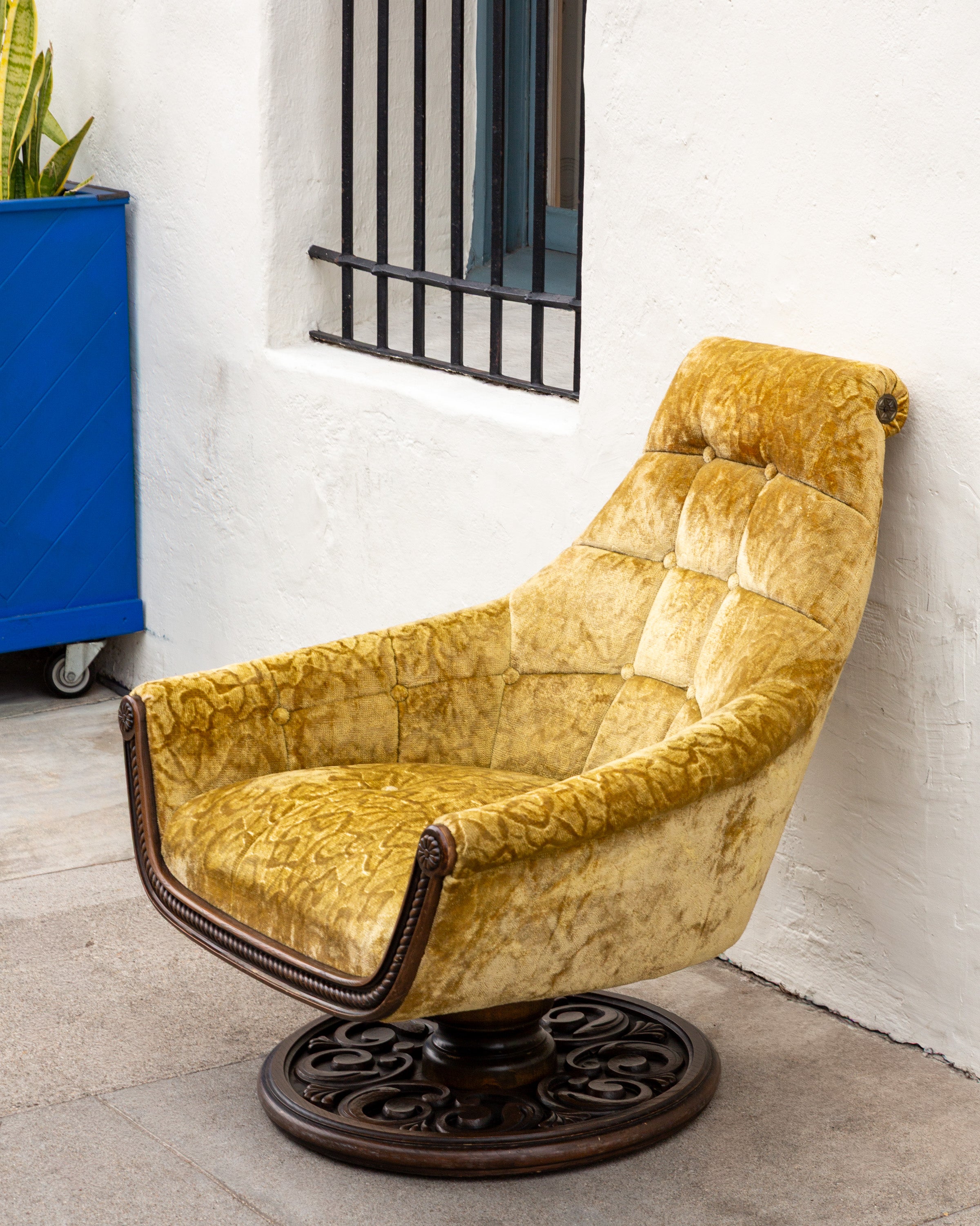 Three-quarter view of a 1970s gold velvet swivel chair on a carved wooden pedestal base, against a white stucco wall beside a blue planter