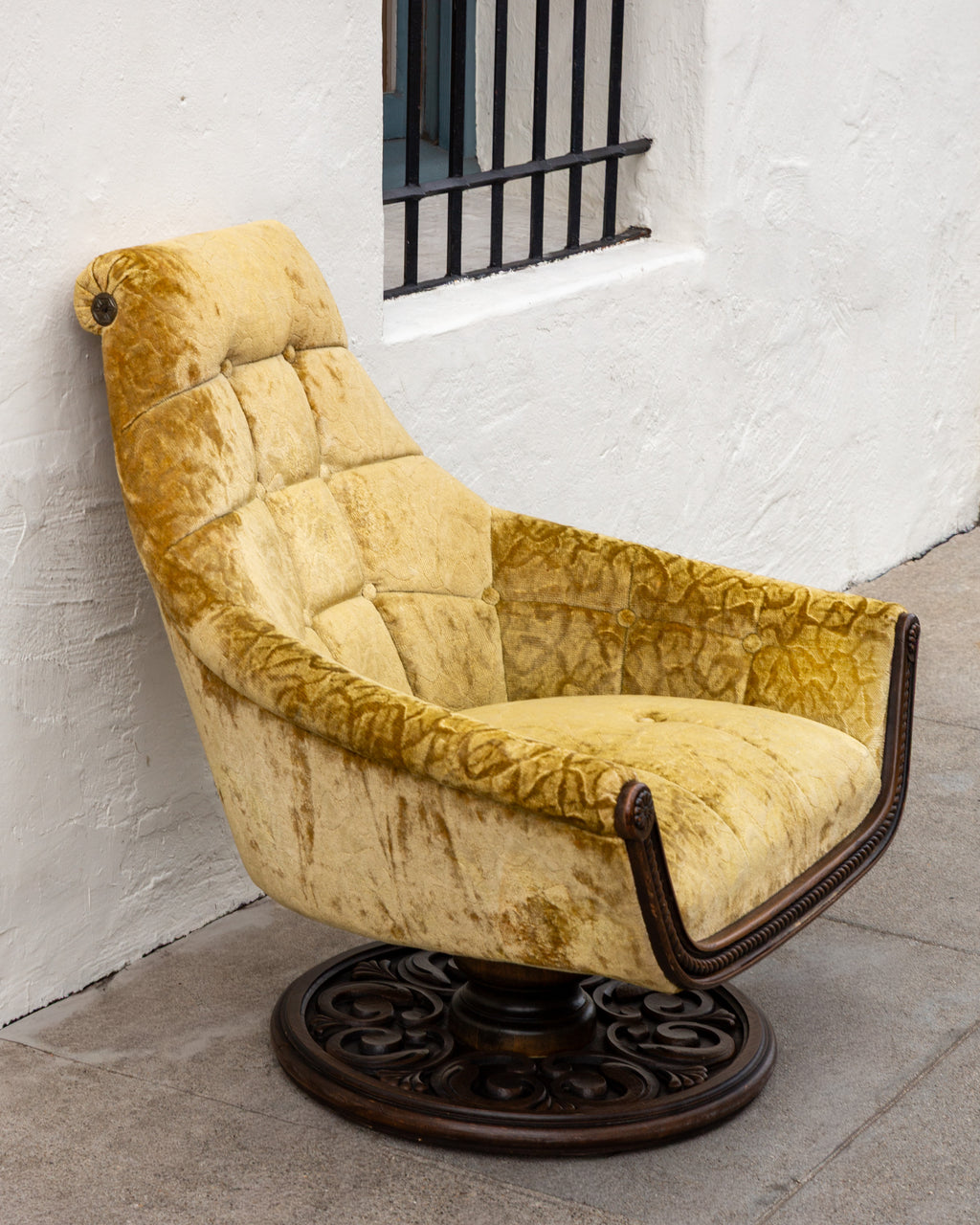 Angled view of the gold velvet swivel chair on the sidewalk, showing the pierced scrollwork carving of the pedestal base