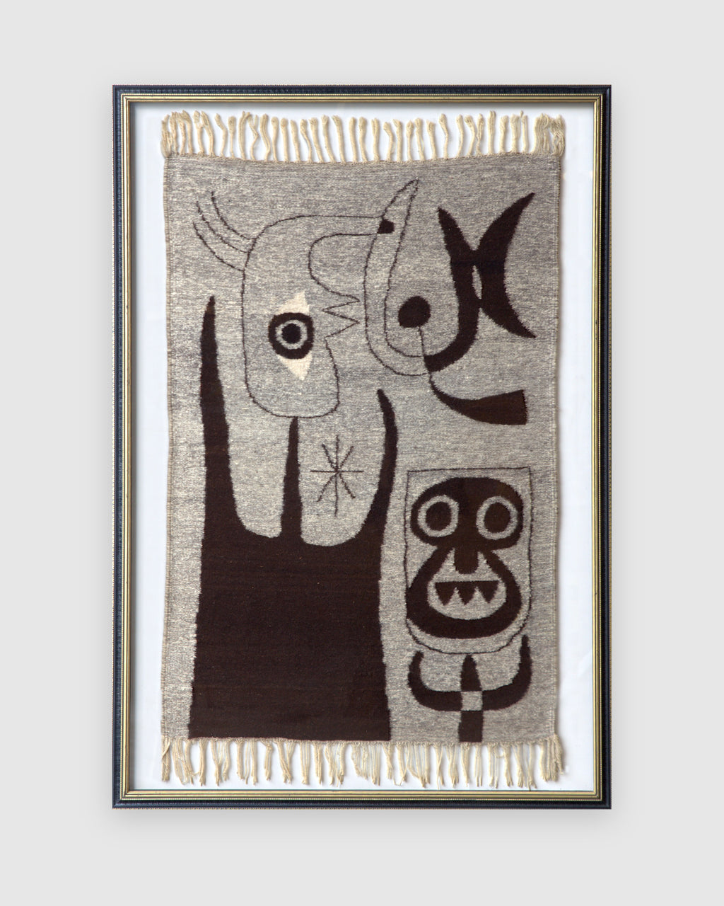 Straight-on view of a framed woven wool tapestry with abstract figures in dark brown on gray, cream fringe top and bottom, in a black and gold frame, on a light gray background.
