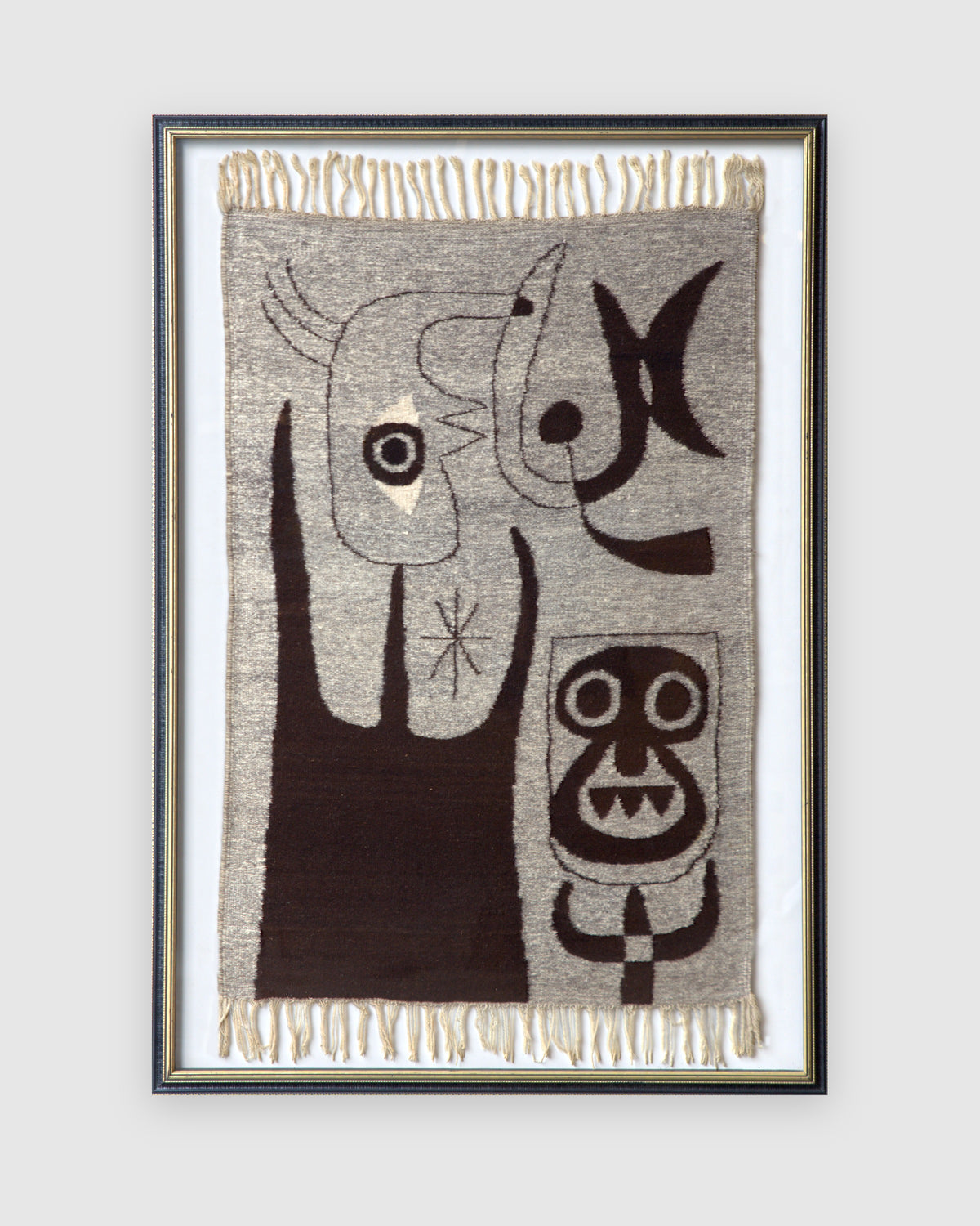 Straight-on view of a framed woven wool tapestry with abstract figures in dark brown on gray, cream fringe top and bottom, in a black and gold frame, on a light gray background.