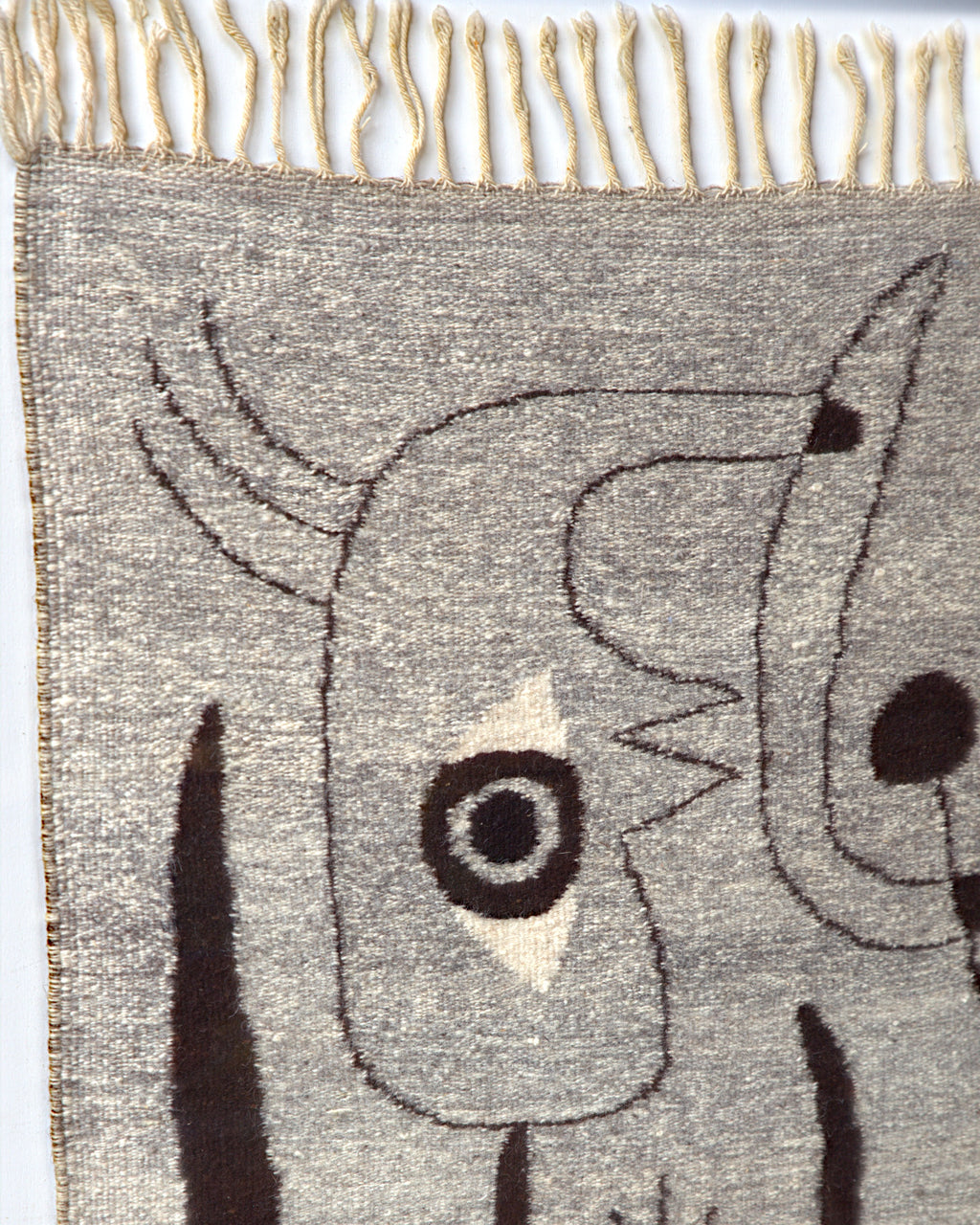 Upper left of the woven tapestry, a bird-like head outlined in dark brown with a ringed eye on a cream diamond, below the cream fringe.