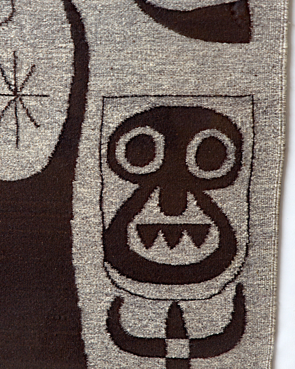 Detail of the mask-like face with round eyes and jagged teeth woven in dark brown on gray wool.