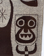 Detail of the mask-like face with round eyes and jagged teeth woven in dark brown on gray wool.