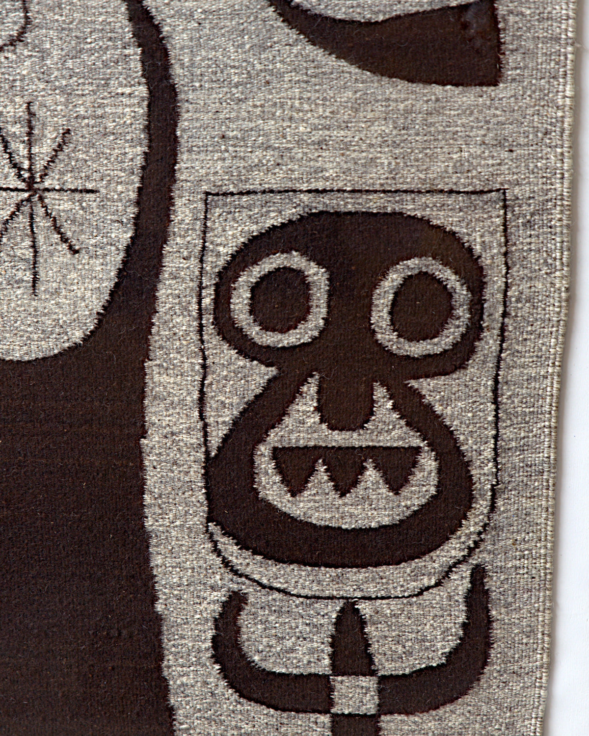 Detail of the mask-like face with round eyes and jagged teeth woven in dark brown on gray wool.