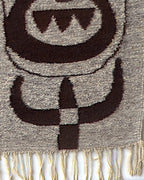 Lower right of the woven tapestry, horned shapes and a checkered stem in dark brown above the cream fringe.