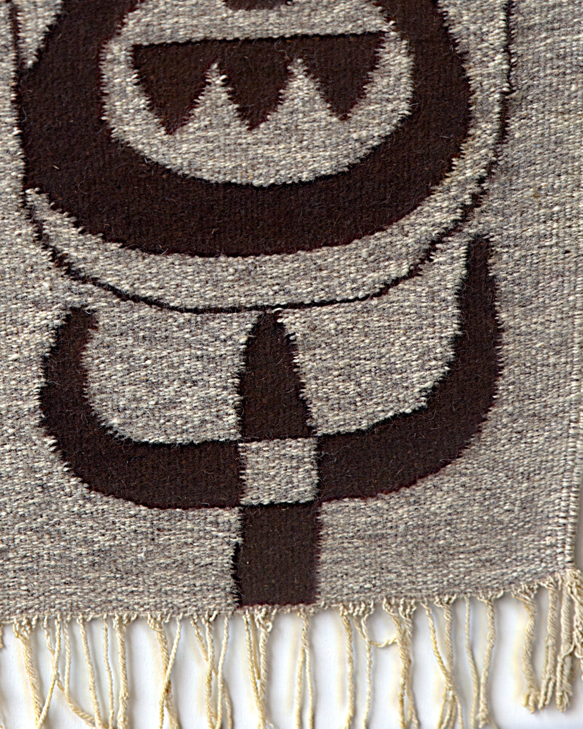 Lower right of the woven tapestry, horned shapes and a checkered stem in dark brown above the cream fringe.